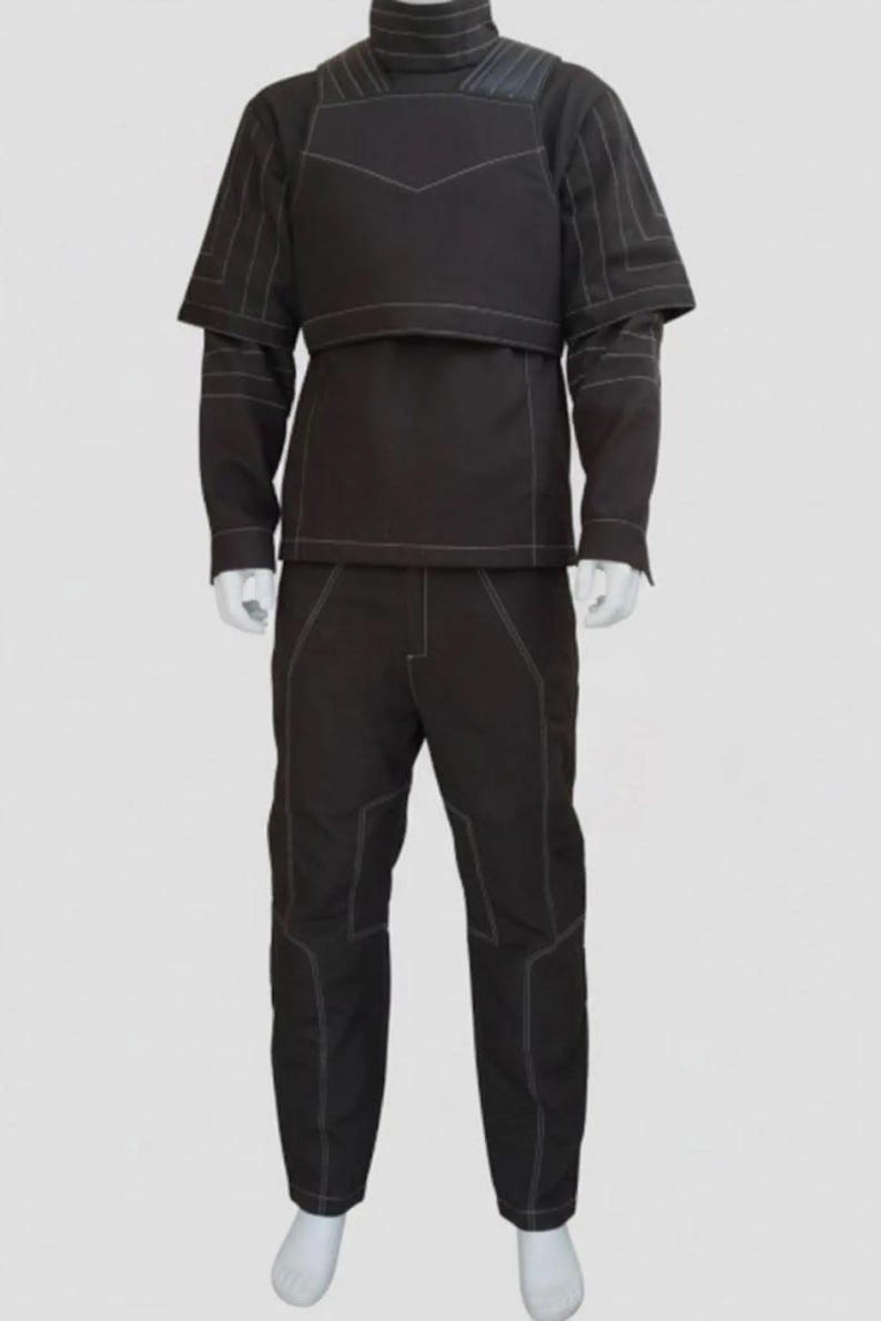 May include: A dark brown, two-piece outfit with white stitching. The top has a high collar and a layered design, with a vest-like overlay. The long-sleeved shirt has a short cape-like detail. The pants are straight-legged. The outfit is displayed on a mannequin.