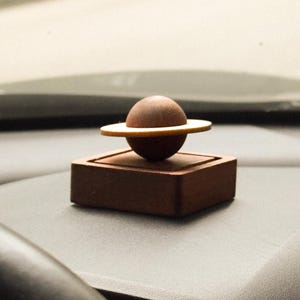 May include: A dark brown wooden object featuring a spherical centre piece encircled by a flat, thin ring, all mounted on a square, solid wooden base. The design resembles a planet with rings, crafted from smooth, matte-finished wood components.