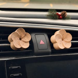 May include: Two light brown wooden flower-shaped air freshener clips attached to the horizontal slats of a car dashboard air vent. The wooden flowers feature five rounded petals and a central detail with thin lines resembling stamens.