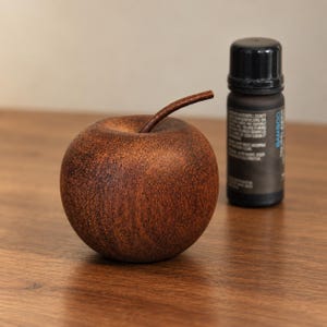 Peut inclure: Une sculpture de pomme en bois rond de couleur brun foncé avec une tige incurvée, placée à côté d'une petite bouteille cylindrique noire avec un bouchon noir et une étiquette portant le mot Bamboo en texte bleu vertical sur le côté.