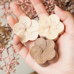 Peut inclure: Trois objets en bois en forme de fleur, dans des tons de marron clair et foncé. Chaque fleur présente un motif central détaillé avec des lignes et des points gravés. Les objets sont tenus dans une main, avec un arrière-plan floral flou.