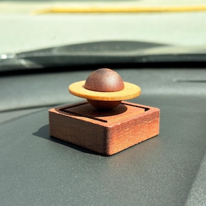 May include: A wooden decorative object resembling a planet. The design features a dark brown sphere atop a light brown ring, set on a square, reddish-brown base. The object is crafted from wood and has a smooth finish.