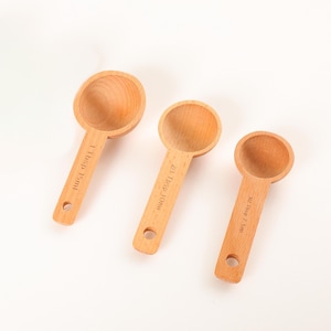 Peut inclure: Trois cuillères à mesurer en bois clair avec des mesures gravées. Les cuillères ont une forme de bol arrondie. Les manches indiquent les mesures en cuillères à soupe et en millilitres.