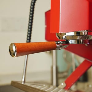 May include: Close-up of a red espresso machine with a wooden handle. The handle has a silver metal cap on the end. The machine has a silver portafilter and a black hose.