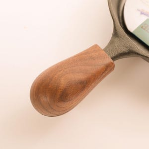 May include: A cast iron skillet handle featuring a smooth, polished brown wooden grip with visible grain patterns. The handle is securely attached to the dark, textured metal body of the skillet, which shows a partial paper label with visible text.