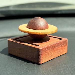 May include: A wooden object featuring a dark brown spherical top piece resting on a flat, light-colored circular disc, which is positioned atop a dark brown square base. The entire structure is crafted from wood with visible grain textures.