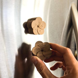 Peut inclure: Deux aimants en bois en forme de fleur avec des détails de pétales gravés. Un aimant est en bois clair fixé sur une surface métallique, tandis que l'autre est en bois plus foncé, montrant le design floral à cinq pétales et le centre.