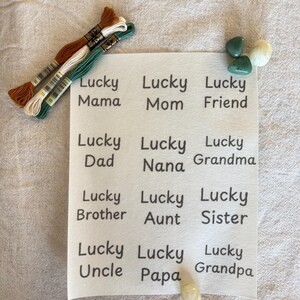 以下が含まれることがあります： 「Lucky」と家族の名前が黒で印刷された白い長方形のカード。茶色、白、緑の刺繍糸が3つ左側にあります。緑と白の石が右側にあります。