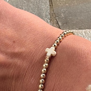 Op de afbeelding: Gouden kralenarmband met een witte kruisbedel. De armband heeft kleine, ronde gouden kralen, met een enkele, gebroken witte kruisbedel in het midden. De armband wordt om een pols getoond.