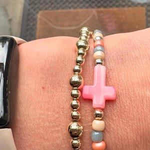May include: Two beaded bracelets. One is gold-coloured with round beads. The other has a pink cross charm and beads in pink, grey, and peach. The bracelets are worn on a wrist with a smartwatch.