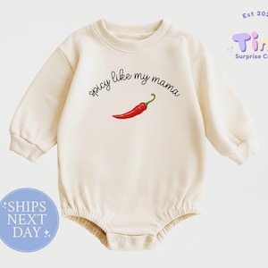 May include: A cream-colored long-sleeve baby romper featuring a crew neckline and ribbed cuffs. The center of the chest displays the black text "spicy like my mama" arched above a single, detailed illustration of a bright red chili pepper.