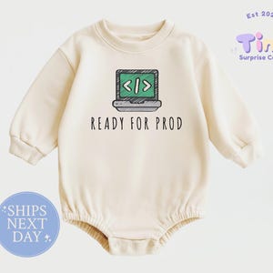 May include: A cream-colored long-sleeve baby romper featuring a graphic of a laptop with a green screen displaying code brackets. Below the laptop, the text reads READY FOR PROD in a simple black font. The garment has a crew neckline and ribbed cuffs.