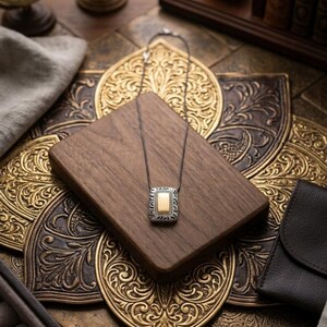 Puede incluir: Un collar de plata con un colgante rectangular con un centro dorado y detalles plateados ornamentados. El colgante descansa sobre un bloque de madera oscura, sobre un fondo de metal dorado intrincadamente tallado. Se ve un estuche de cuero en el fondo.