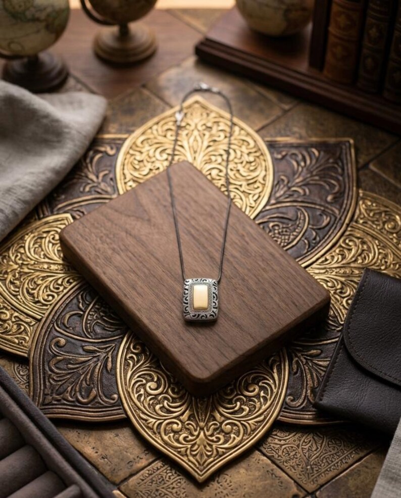 Puede incluir: Un collar de plata con un colgante rectangular con un centro dorado y detalles plateados ornamentados. El colgante descansa sobre un bloque de madera oscura, sobre un fondo de metal dorado intrincadamente tallado. Se ve un estuche de cuero en el fondo.