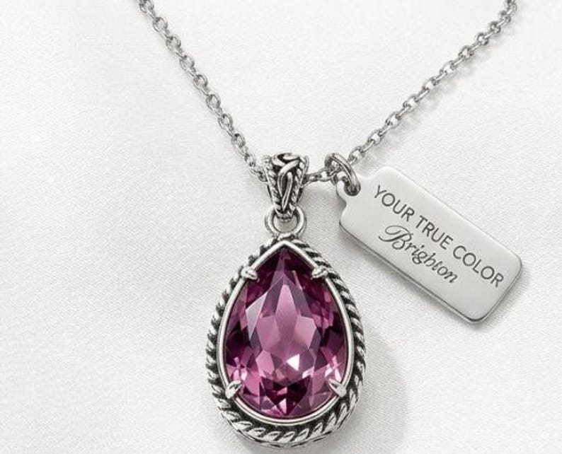 Puede incluir: Collar plateado con un colgante grande en forma de l&aacute;grima. El colgante presenta una piedra preciosa facetada de color rosa intenso, enmarcada por un dise&ntilde;o de cuerda trenzada. Una peque&ntilde;a etiqueta con el texto "YOUR TRUE COLOR Brighton" est&aacute; unida a la cadena.
