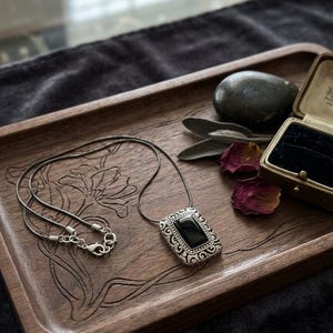 Puede incluir: Un collar de plata con un colgante rectangular de piedra negra descansa sobre una bandeja de madera con tallas florales. El colgante tiene un borde plateado adornado. Una peque&ntilde;a caja de joyas y una piedra oscura tambi&eacute;n est&aacute;n en la bandeja.