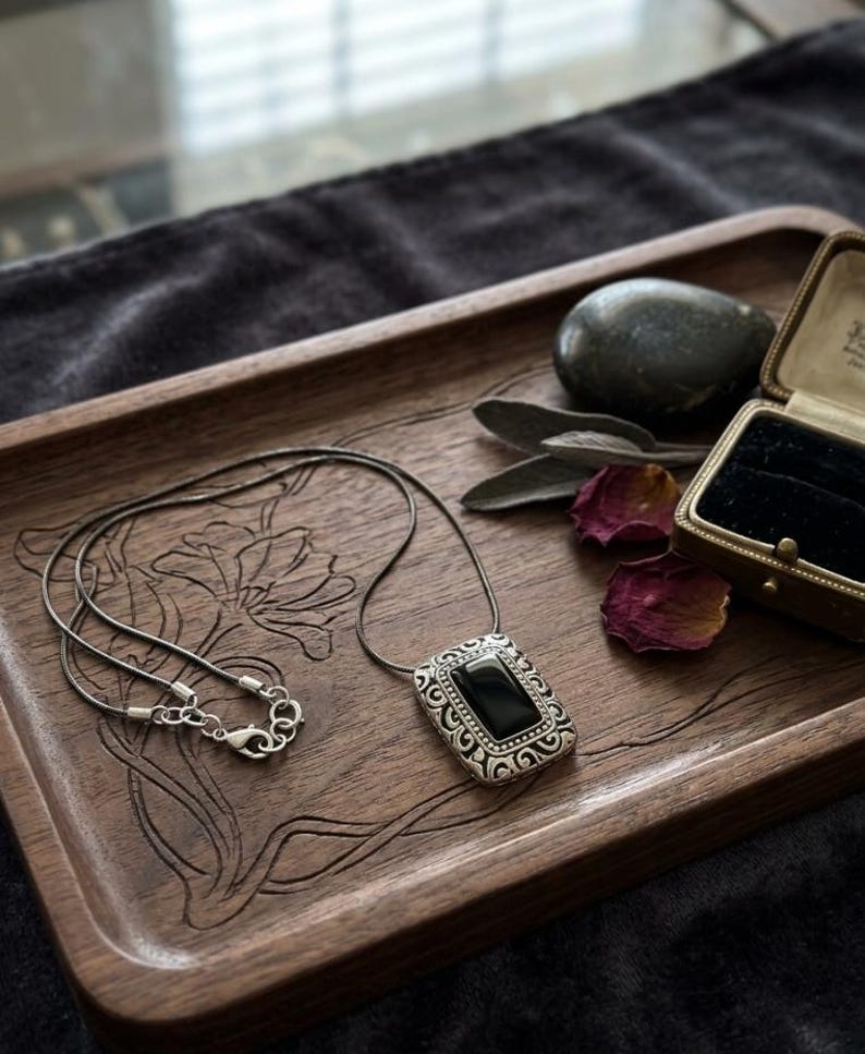 Puede incluir: Un collar de plata con un colgante rectangular de piedra negra descansa sobre una bandeja de madera con tallas florales. El colgante tiene un borde plateado adornado. Una peque&ntilde;a caja de joyas y una piedra oscura tambi&eacute;n est&aacute;n en la bandeja.