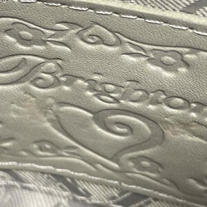 May include: A light gray leather patch featuring the embossed word Brighton in a stylized script font. The patch is decorated with an embossed floral pattern and a heart shape surrounding the brand name, with visible stitching along the top edge.
