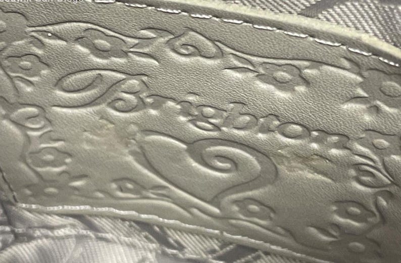 May include: A light gray leather patch featuring the embossed word Brighton in a stylized script font. The patch is decorated with an embossed floral pattern and a heart shape surrounding the brand name, with visible stitching along the top edge.
