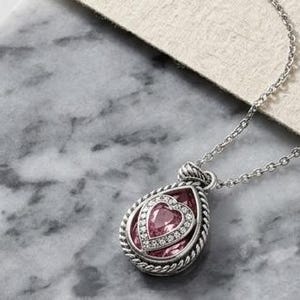 Puede incluir: Un collar plateado con un colgante en forma de l&aacute;grima. El colgante presenta una piedra rosa en forma de coraz&oacute;n, rodeada por un halo de peque&ntilde;as piedras transparentes y un borde plateado trenzado. La cadena tambi&eacute;n es plateada.