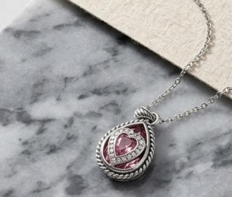 Puede incluir: Un collar plateado con un colgante en forma de l&aacute;grima. El colgante presenta una piedra rosa en forma de coraz&oacute;n, rodeada por un halo de peque&ntilde;as piedras transparentes y un borde plateado trenzado. La cadena tambi&eacute;n es plateada.