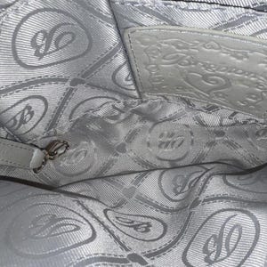 May include: The interior of a light gray handbag featuring a repeating pattern of the letter B and heart shapes on the fabric lining. A rectangular leather patch embossed with the word Brighton and a heart design is stitched to the upper back wall.