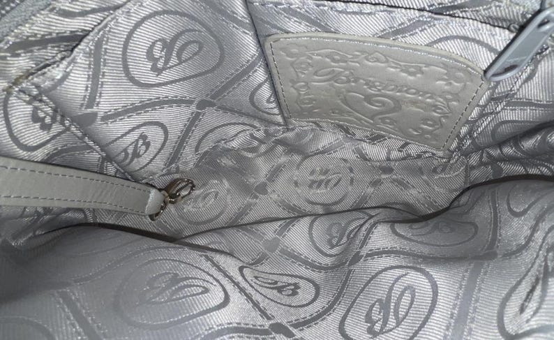 May include: The interior of a light gray handbag featuring a repeating pattern of the letter B and heart shapes on the fabric lining. A rectangular leather patch embossed with the word Brighton and a heart design is stitched to the upper back wall.