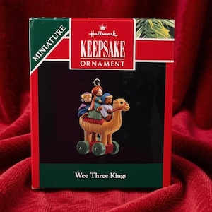 May include: A Hallmark Keepsake miniature ornament box featuring the text Wee Three Kings. The box displays a graphic of three figures riding a brown camel on wheels. The packaging is red, green, and black with a gold hanging loop at the top.