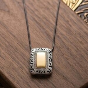 Puede incluir: Un collar plateado con un colgante rectangular. El colgante tiene un centro dorado y un borde plateado decorativo. El collar se muestra sobre una superficie de madera.