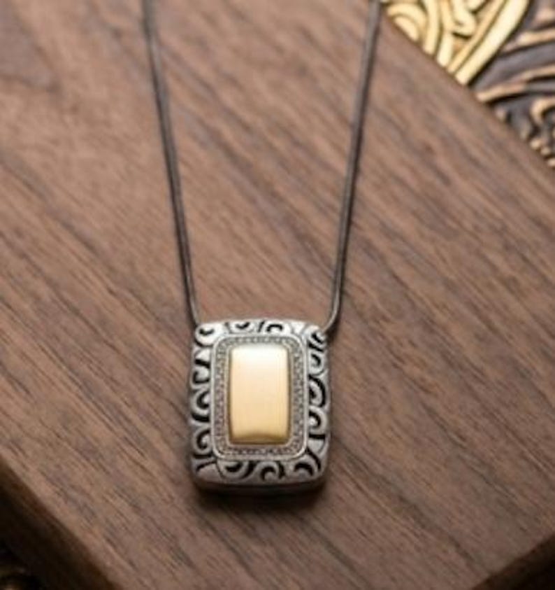 Puede incluir: Un collar plateado con un colgante rectangular. El colgante tiene un centro dorado y un borde plateado decorativo. El collar se muestra sobre una superficie de madera.