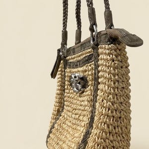 May include: A beige woven straw handbag featuring dark brown braided shoulder straps and dark brown snakeskin-textured trim. The bag includes a silver-toned metal zipper closure at the top and a prominent silver-toned decorative emblem on the front.