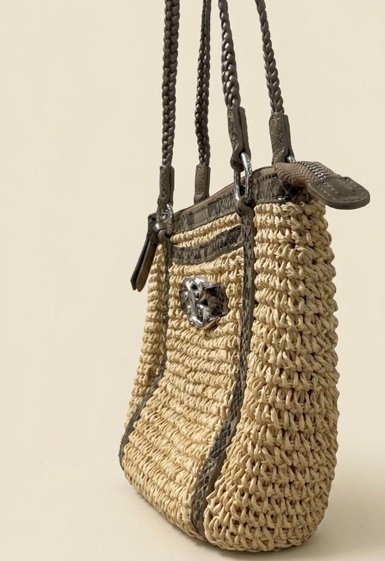 May include: A beige woven straw handbag featuring dark brown braided shoulder straps and dark brown snakeskin-textured trim. The bag includes a silver-toned metal zipper closure at the top and a prominent silver-toned decorative emblem on the front.