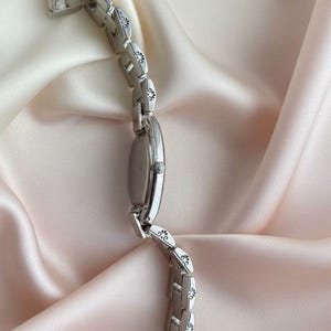 May include: A silver-toned wristwatch featuring an oval-shaped case and a link bracelet. Each individual link of the bracelet is decorated with a small, engraved black floral or scroll pattern. The watch includes a side crown for time adjustment.