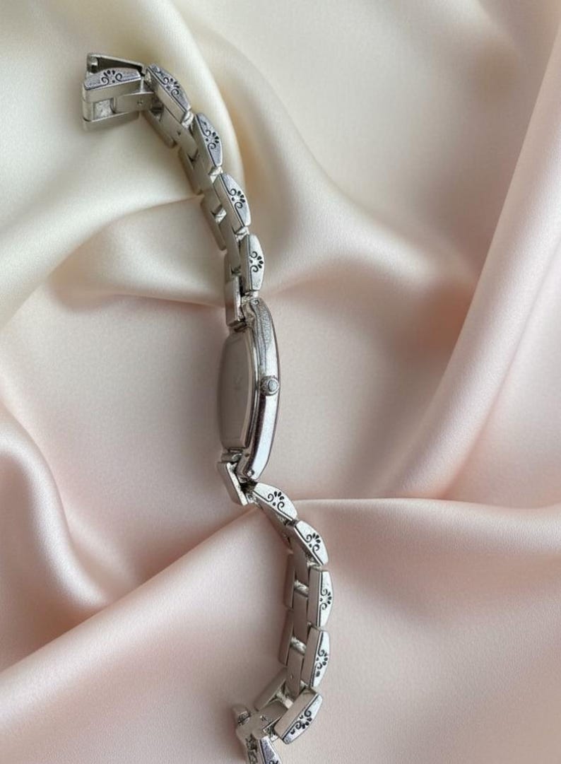 May include: A silver-toned wristwatch featuring an oval-shaped case and a link bracelet. Each individual link of the bracelet is decorated with a small, engraved black floral or scroll pattern. The watch includes a side crown for time adjustment.