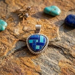 Puede incluir: Un colgante triangular con una incrustación geométrica de segmentos de lapislázuli azul oscuro y turquesa azul claro. El colgante está enmarcado por un borde de cuerda de plata trenzada e incluye una anilla de plata en la parte superior.