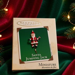 Może przedstawiać: Pudełko na ozdobę Hallmark Keepsake z miniaturką Santa Jumping Jack. Pudełko jest kremowe z zielonym panelem środkowym, na którym znajduje się figurka Świętego Mikołaja trzymająca laskę cukrową i zielony worek, z napisem Santa Jumping Jack.