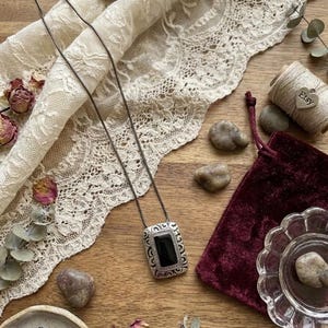 Puede incluir: Un collar con colgante rectangular plateado con un centro de piedra negra, exhibido sobre una superficie de madera con encaje, flores secas y una bolsa de terciopelo. El colgante tiene un borde ornamentado y detallado.