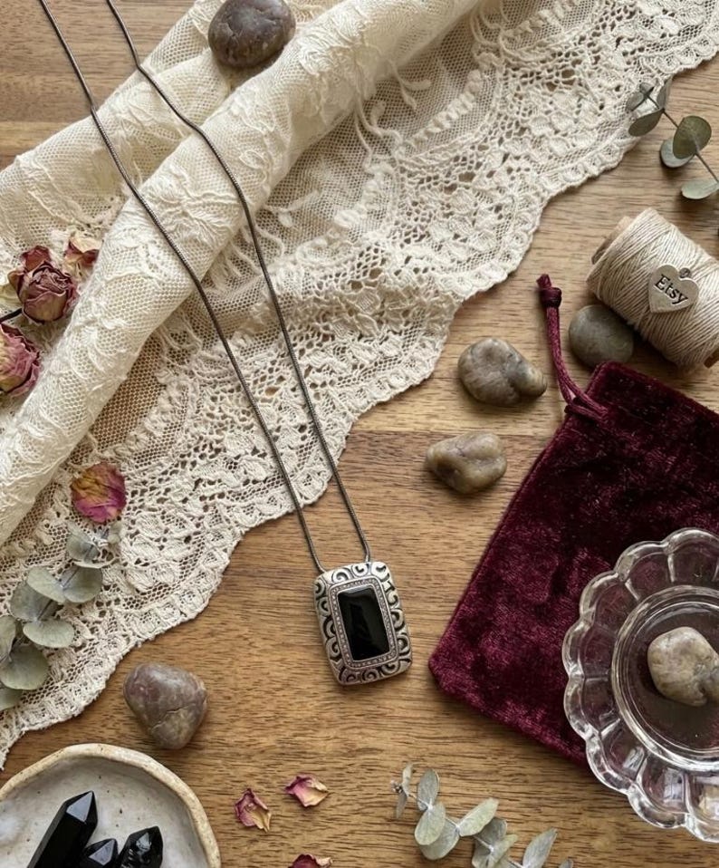 Puede incluir: Un collar con colgante rectangular plateado con un centro de piedra negra, exhibido sobre una superficie de madera con encaje, flores secas y una bolsa de terciopelo. El colgante tiene un borde ornamentado y detallado.