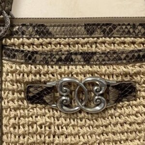 May include: A beige woven straw handbag featuring brown and black snakeskin-patterned leather trim, silver-toned metal hardware, and a central decorative silver emblem with an intricate swirling design attached to the front panel.