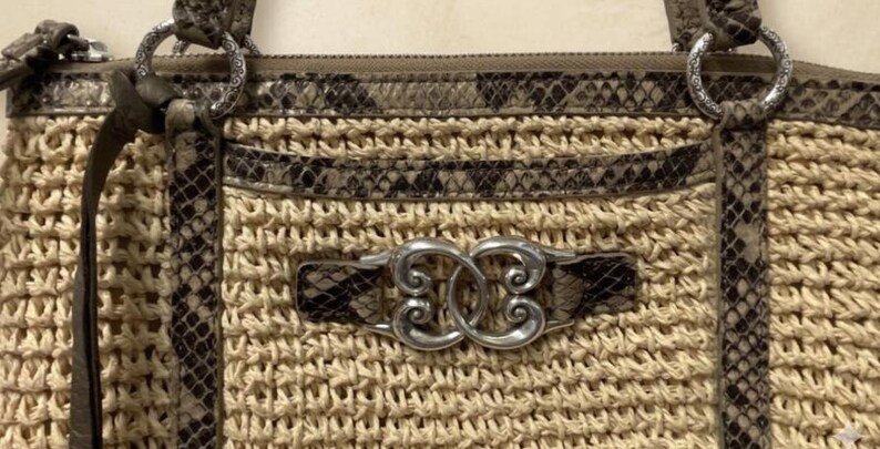 May include: A beige woven straw handbag featuring brown and black snakeskin-patterned leather trim, silver-toned metal hardware, and a central decorative silver emblem with an intricate swirling design attached to the front panel.