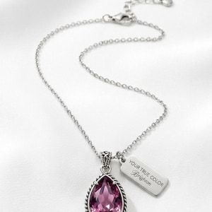 Puede incluir: Collar plateado con un colgante rosa facetado en forma de l&aacute;grima. El colgante tiene un borde trenzado. Una peque&ntilde;a etiqueta rectangular con el texto "YOUR TRUE COLOR Brighton" cuelga junto al colgante.