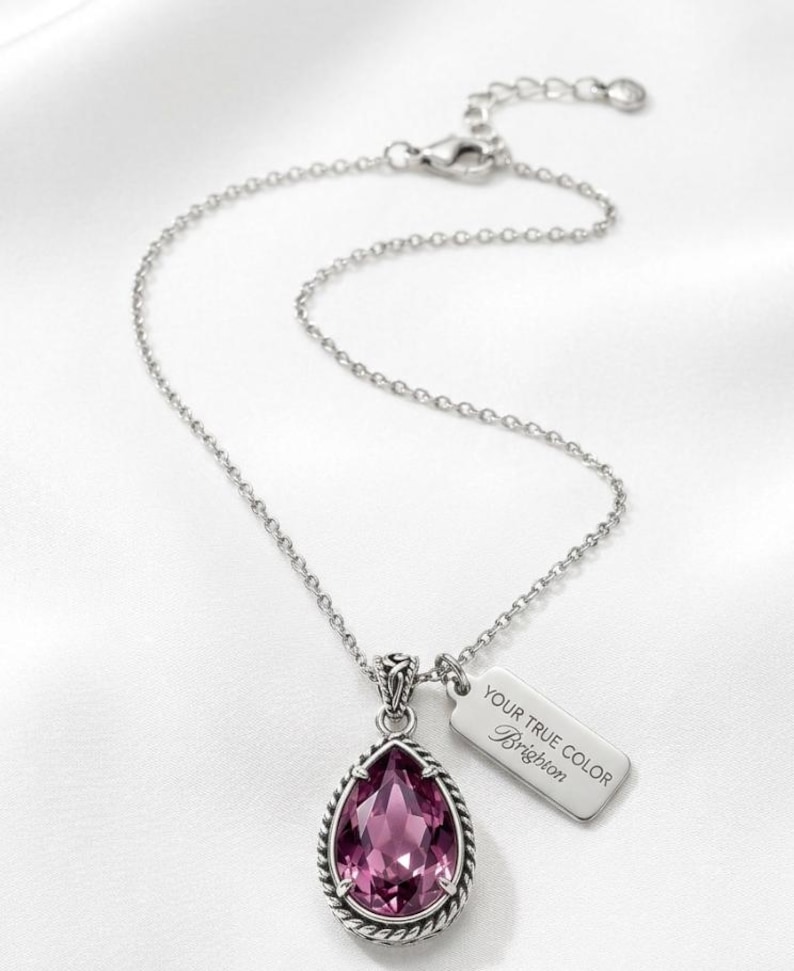Puede incluir: Collar plateado con un colgante rosa facetado en forma de l&aacute;grima. El colgante tiene un borde trenzado. Una peque&ntilde;a etiqueta rectangular con el texto "YOUR TRUE COLOR Brighton" cuelga junto al colgante.