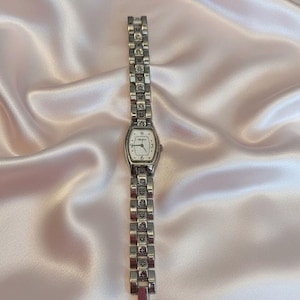 May include: A silver-tone analog wristwatch featuring a rectangular face with a white dial, black numerals at the three, six, and nine o'clock positions, and a decorative link bracelet with intricate engraved patterns on each metal segment.