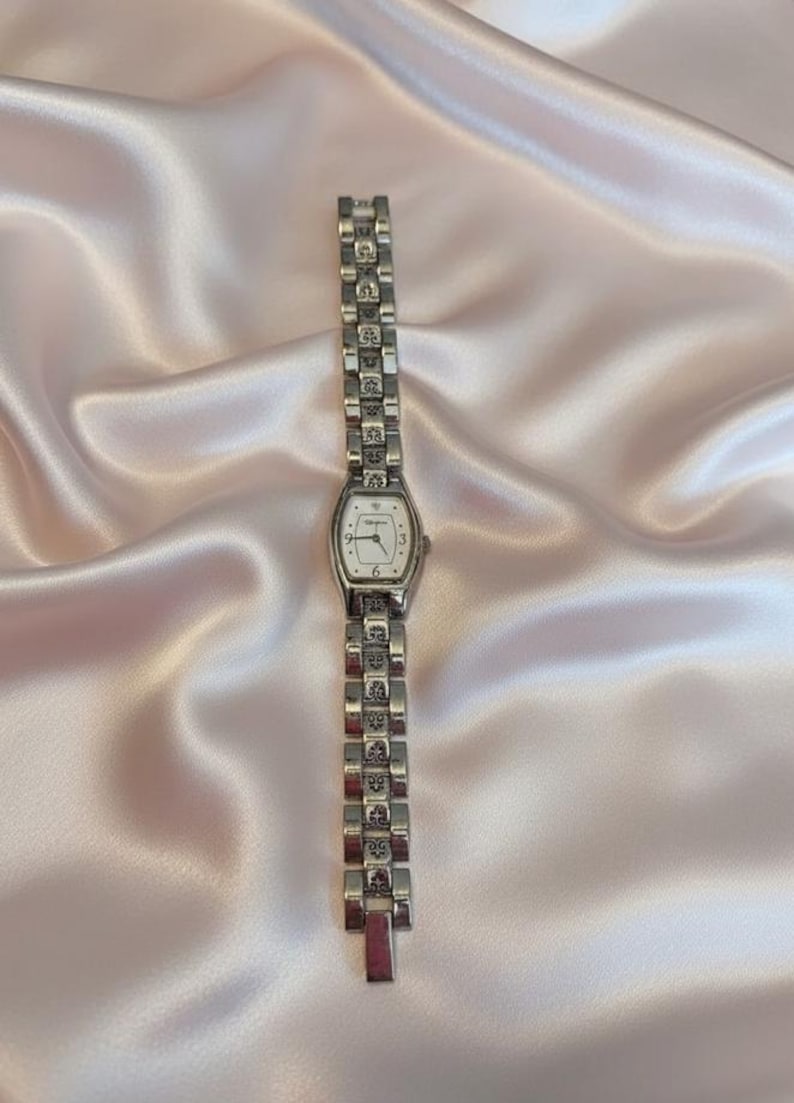 May include: A silver-tone analog wristwatch featuring a rectangular face with a white dial, black numerals at the three, six, and nine o'clock positions, and a decorative link bracelet with intricate engraved patterns on each metal segment.