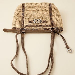 May include: A beige woven straw handbag featuring brown snakeskin-patterned trim, two braided brown leather handles, and a silver-toned metal emblem on the front. A silver heart-shaped charm hangs from the side zipper pull of the bag.
