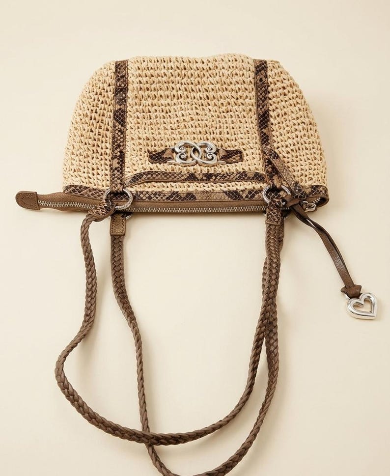 May include: A beige woven straw handbag featuring brown snakeskin-patterned trim, two braided brown leather handles, and a silver-toned metal emblem on the front. A silver heart-shaped charm hangs from the side zipper pull of the bag.
