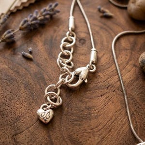 Puede incluir: Primer plano de un collar plateado con un dije en forma de coraz&oacute;n. El dije tiene las iniciales "JB" grabadas. El collar est&aacute; sobre una superficie de madera con lavanda seca y peque&ntilde;as piedras.