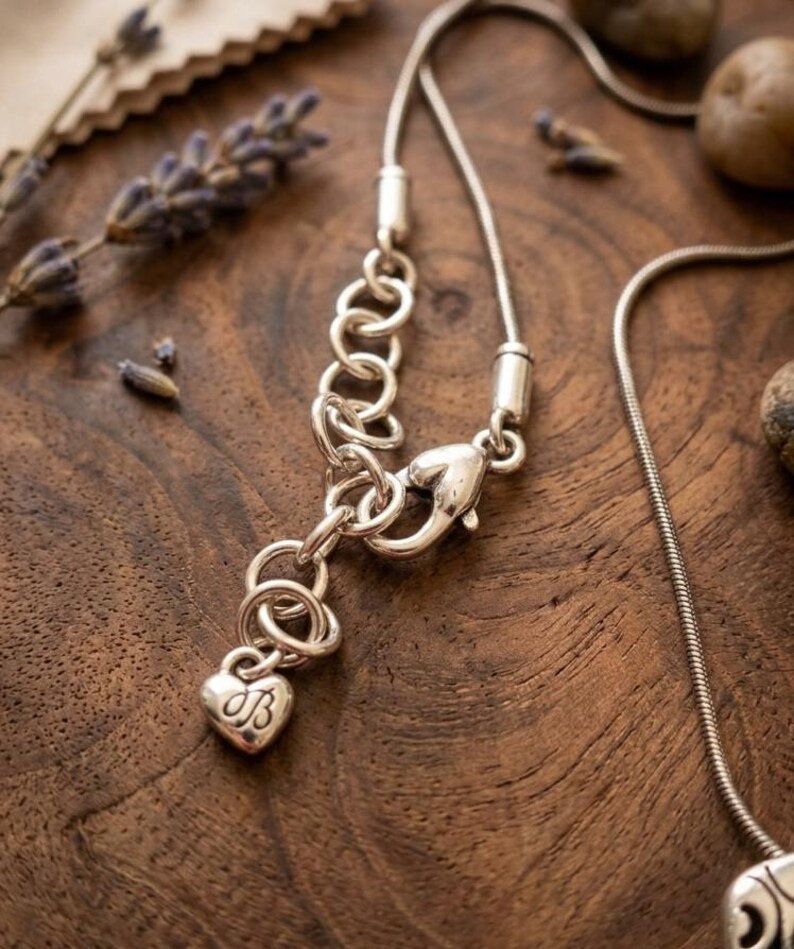 Puede incluir: Primer plano de un collar plateado con un dije en forma de coraz&oacute;n. El dije tiene las iniciales "JB" grabadas. El collar est&aacute; sobre una superficie de madera con lavanda seca y peque&ntilde;as piedras.