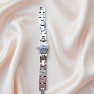 May include: A silver-toned wristwatch featuring a rectangular case with the word Brighton and a small heart symbol engraved on the back. The watch has a linked metal band with alternating plain silver segments and textured pink segments.