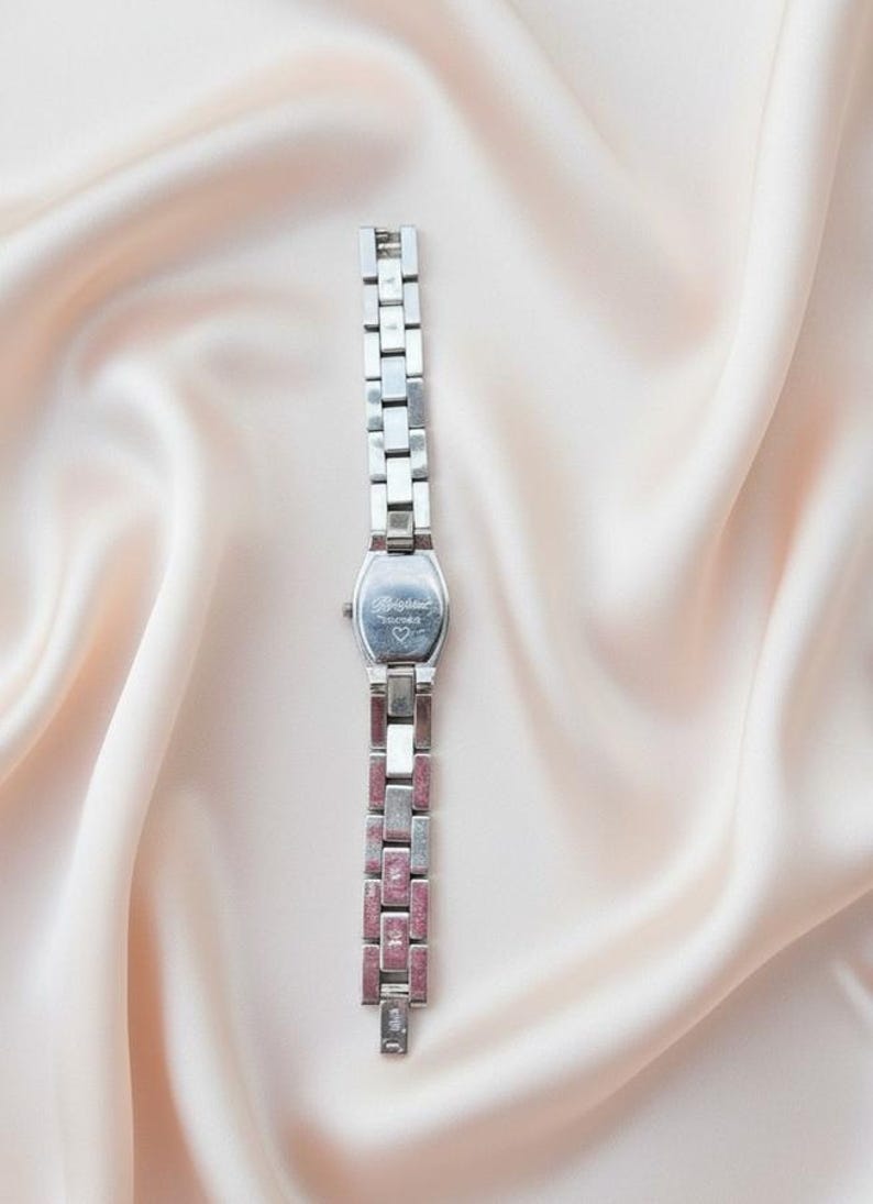 May include: A silver-toned wristwatch featuring a rectangular case with the word Brighton and a small heart symbol engraved on the back. The watch has a linked metal band with alternating plain silver segments and textured pink segments.
